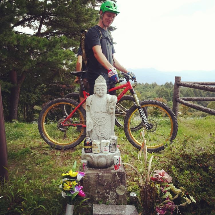 Right side of a cyclist, mounted over a red Surly bike, behind a small Buddha statue with candles and flowers below it