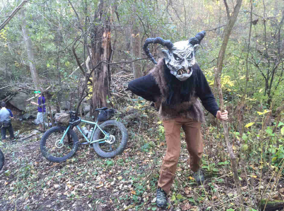 Front view of a person wearing a scary mask with horns, and a fat bike, trees and people in the background