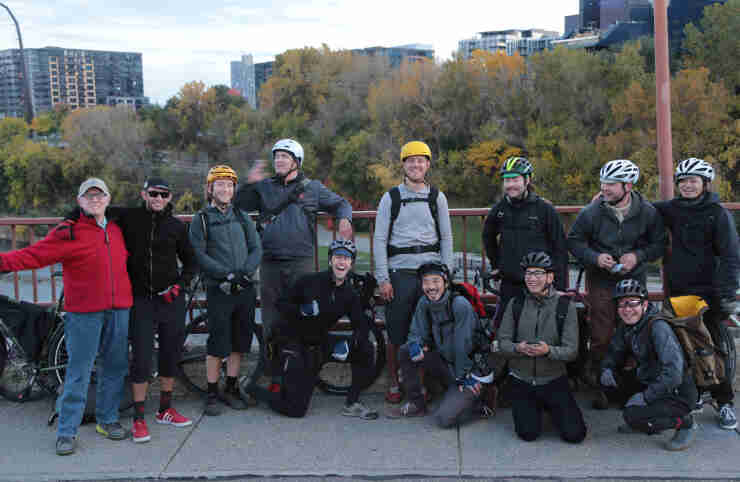 Front view of a group of cyclists posing on a sidewalk, on a bridge, with trees and buildings in the background