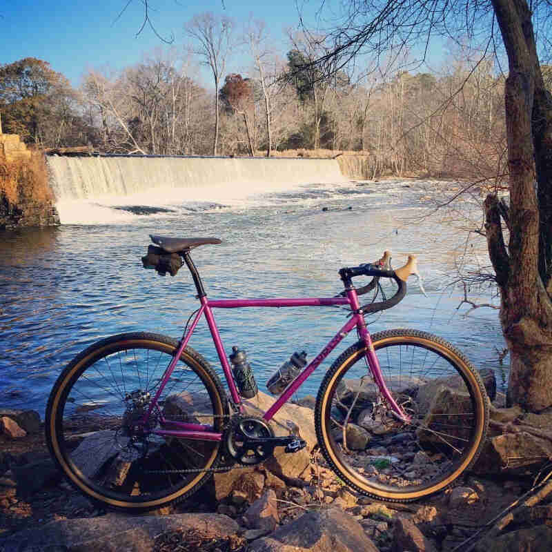 Right side view of a purple Surly Straggler bike, parked on top of a rocky river bank, with a waterfall in background