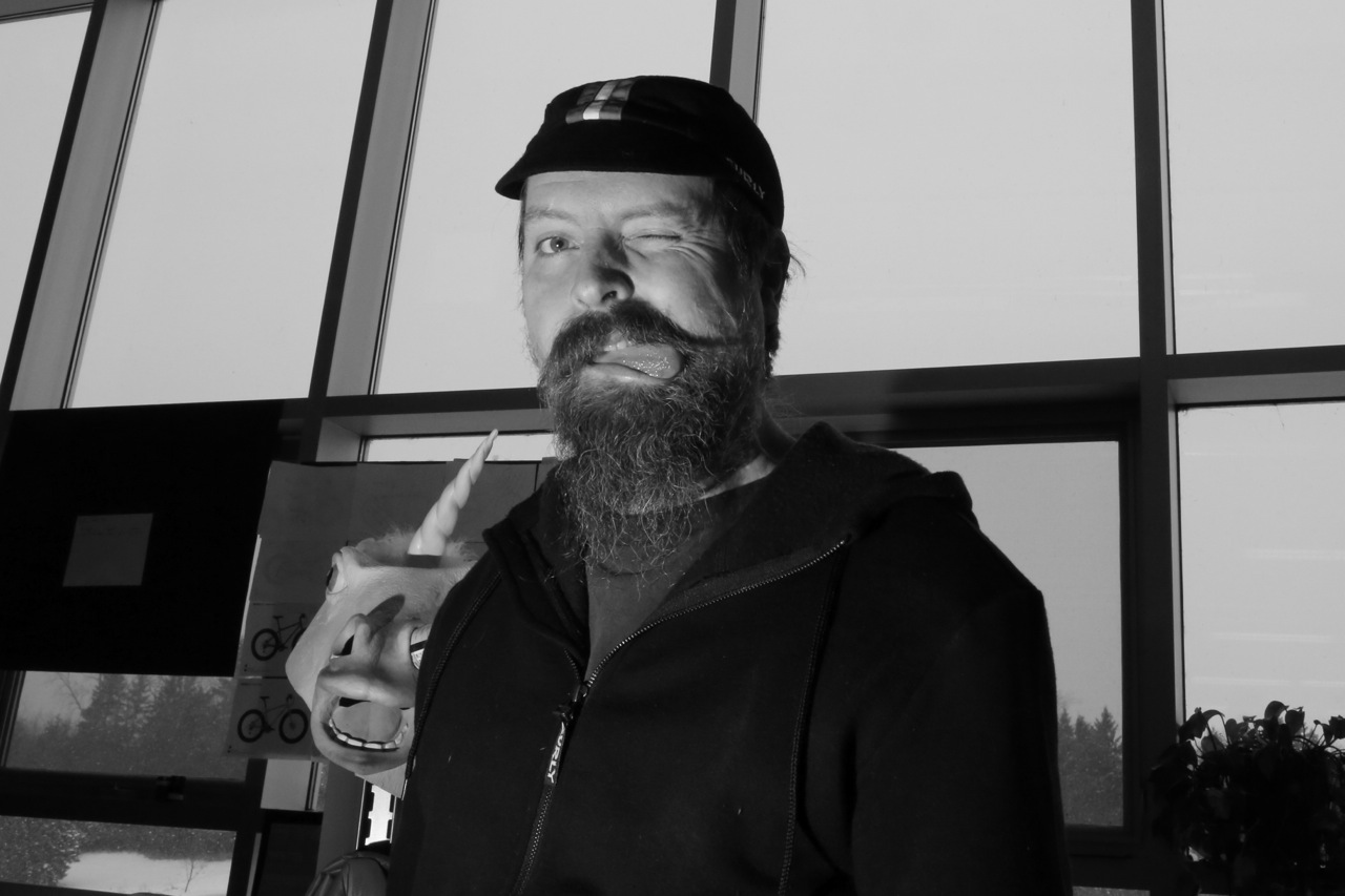 Front view of a person with a beard, biting their tongue, in a room with a glass wall behind them - black & white image