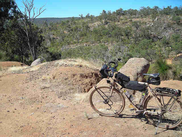 Left side view of a tan Surly Long Haul Trucker bike, parked on a dirt hilltop, with hills and trees in the background