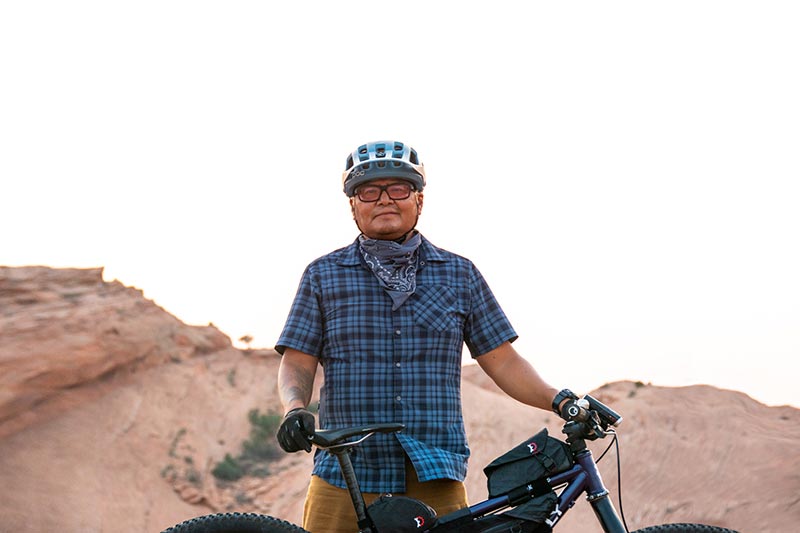 Jon Yazzie standing with loaded mountain bike wearing cycling helmet and riding glasses in front of slick rock