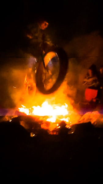 A cyclist riding a fat bike, jumping over a campfire, at night