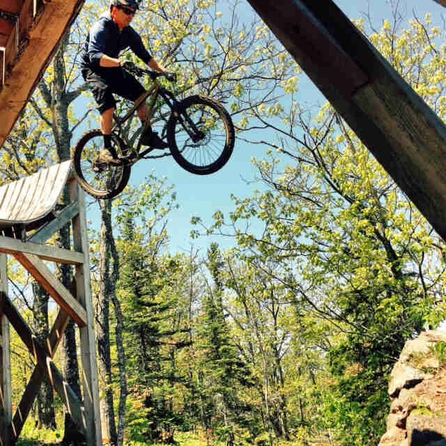 Right side view of a cyclist, on a Surly Instigator bike, flying through the air off of a high ramp in the forest