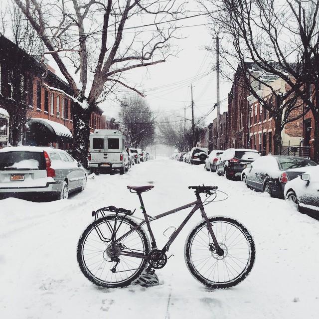 Right side view of a Surly Ogre bike, standing across a snow covered city street with cars parked on a the sides