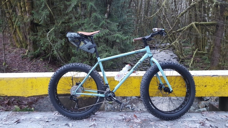 Right side view of a mint Surly Wednesday fat bike, parked along a yellow road barrier, with the woods in the background
