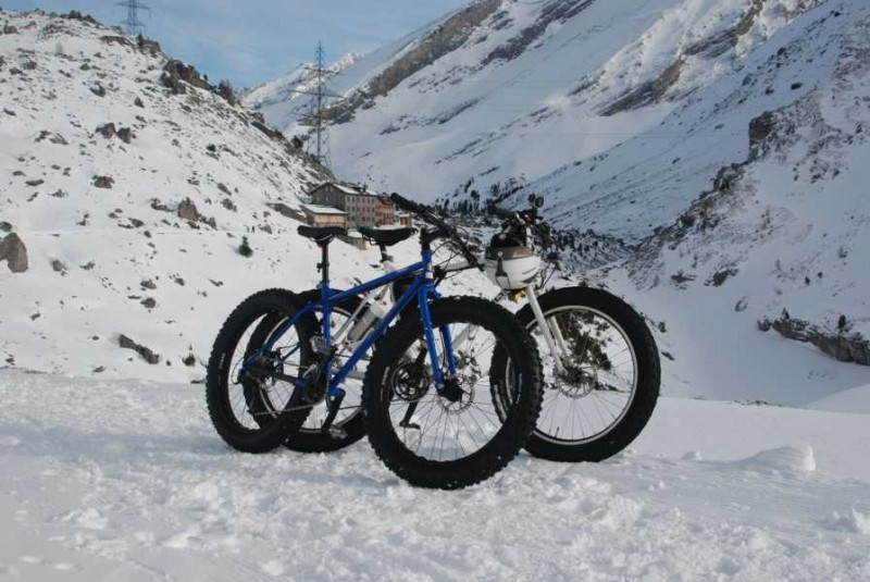 Right side view of 2 Surly fat bikes, parked side by side, on a flat snowy spot below snow capped mountains