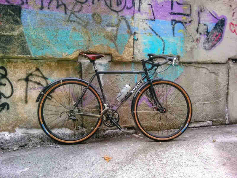 Right side view of a black Surly bike, leaning against a cement wall with graffiti painted on it