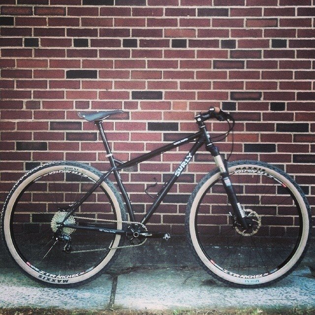 Right side view of a black Surly Karate Monkey bike, parked on a sidewalk, against a brick wall