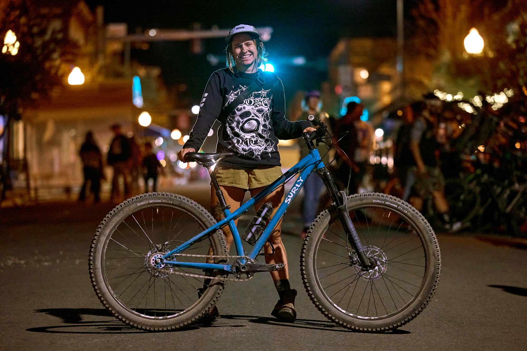 Person standing behind blue single speed Karate Monkey bike in street at night smiling