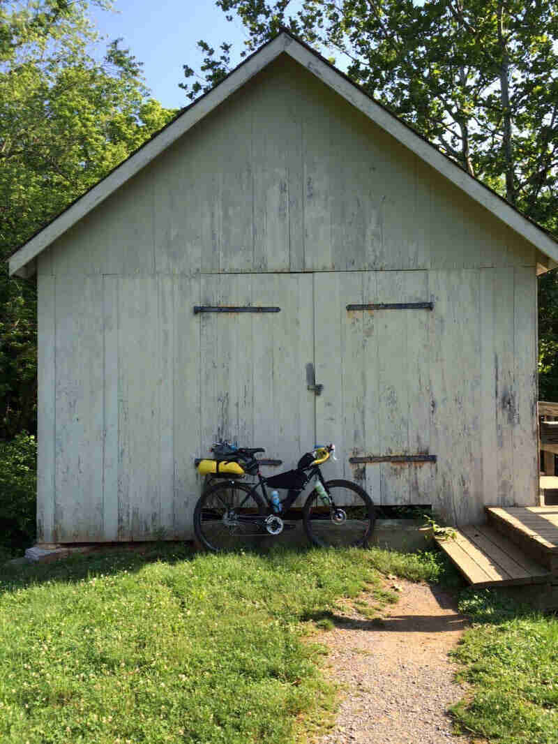 Right side view of a Surly Karate Monkey bike, loaded with gear, leaning on the doors of a white shed with trees behind