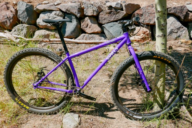 Right side view of a Surly Karate Monkey bike - purple - parked on dirt and weeks - rock wall in the background