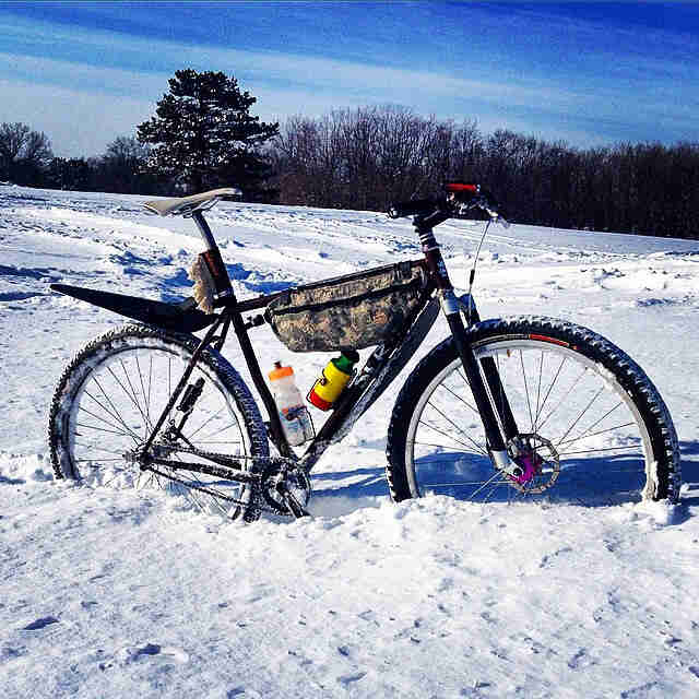 Right side view of a black Surly Karate Monkey bike with frame pack, in a deep snow field with trees in the background