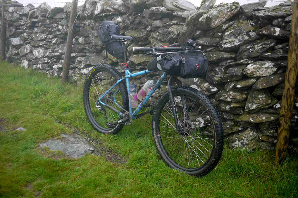 Right side view of a blue Surly Karate Monkey bike, with gear and water bottles, parked in grass along a stone wall