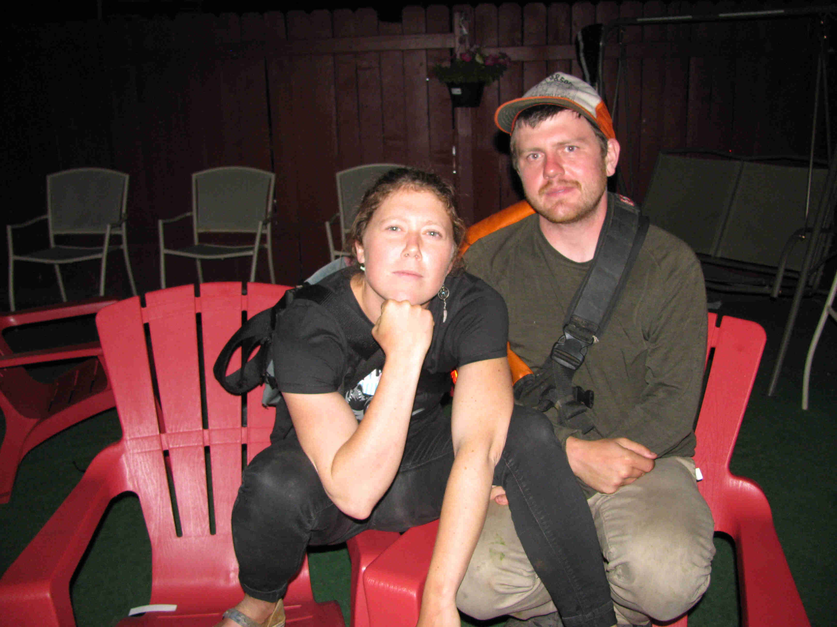 Front view of 2 people sitting on red, plastic chairs, posing together, inside a room with chairs lined up behind them