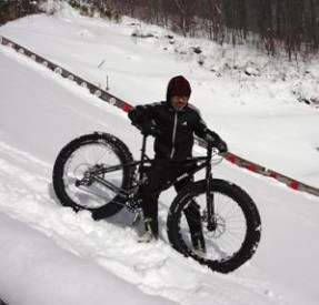 Right side of black Surly fat bike, with a cyclist standing on the left side, sliding down a hill with deep snow on it
