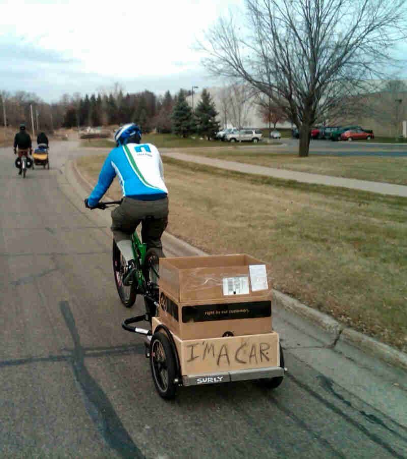 Rear view of a cyclist riding down the side of a street, on a Surly bike with trailer, with a sign showing, I'm a car