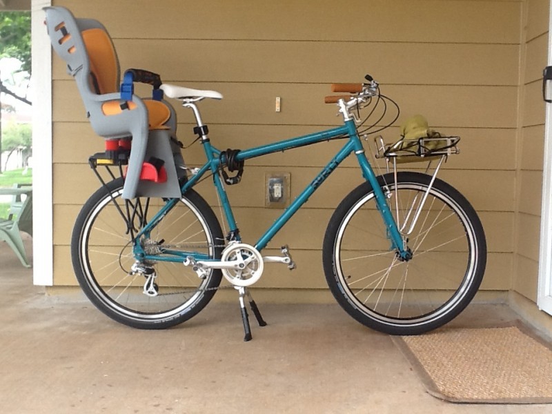 Right side view of a teal Surly Troll bike with a child seat on the back rack, parked on a cement front porch of a house