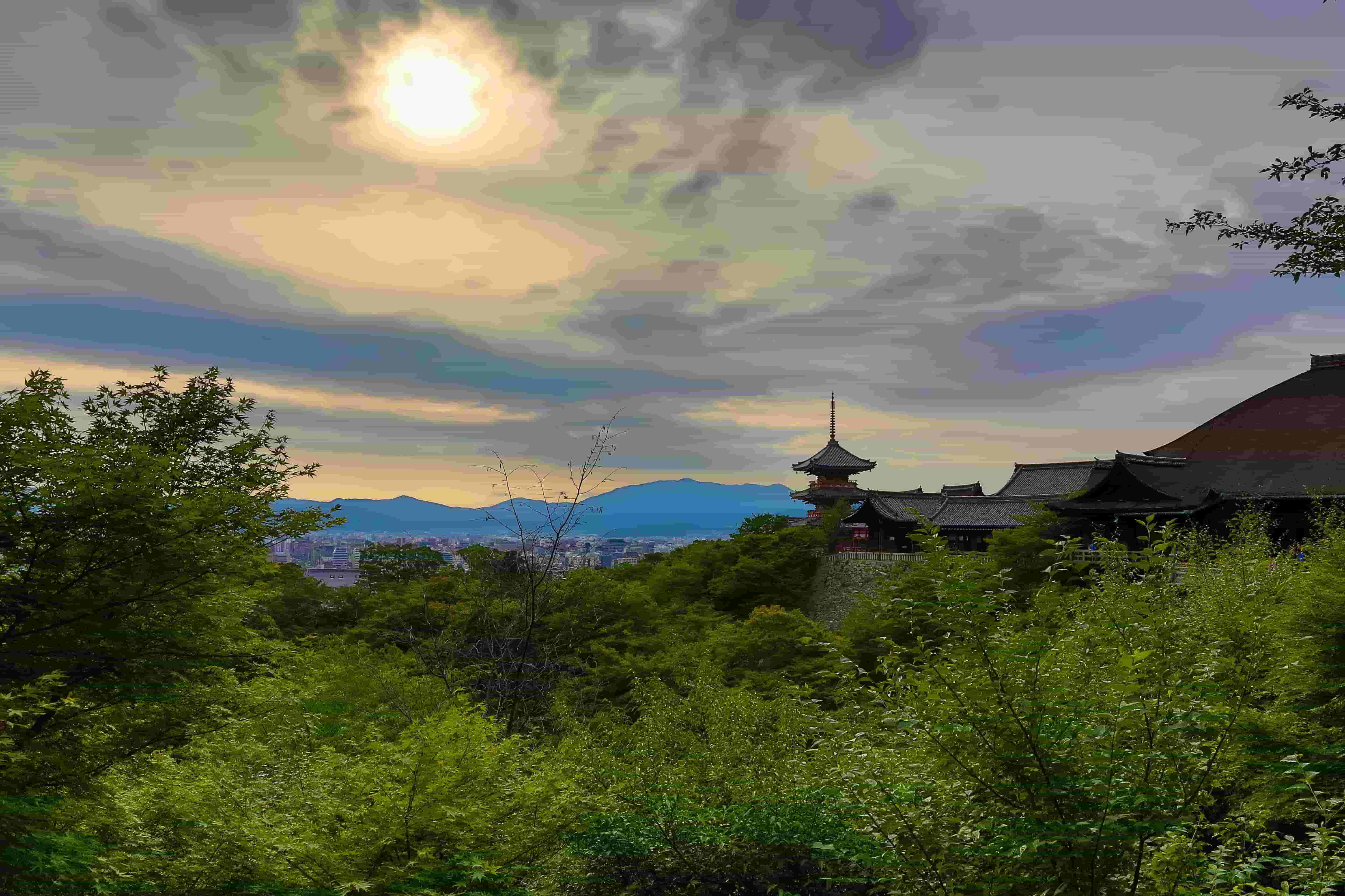 A pagoda roof building, peeking up through tree tops, with a city and hills in the background, and hazy sun above