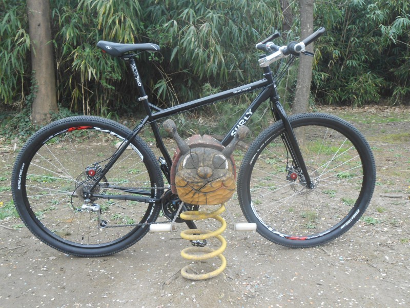Right side view of a black Surly Karate Monkey bike, parked on dirt, with an spring type item in front of the cranks