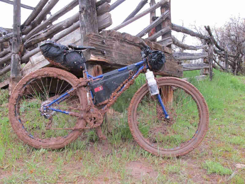 Right side view of a blue Surly Krampus with gear, covered in mud, parked on a grass hill in front of a wood post fence