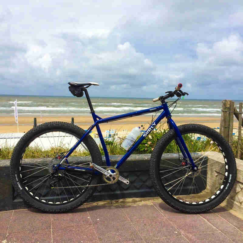 Right side view of a blue Surly Krampus bike, parked on a block patio, with a beach and ocean in the background