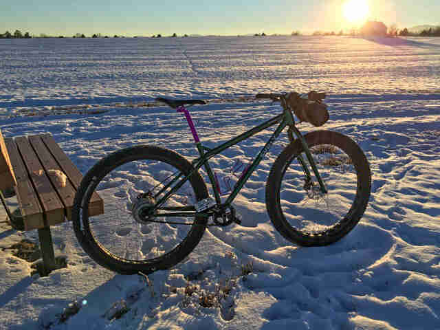 Right side view of a green Surly Krampus bike, parked in a snow covered field, next to a park bench