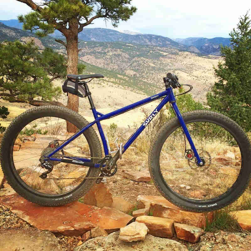 Right side view of a blue Surly Krampus bike, parked on rocky hilltop, with grassy hills and mountains in the background