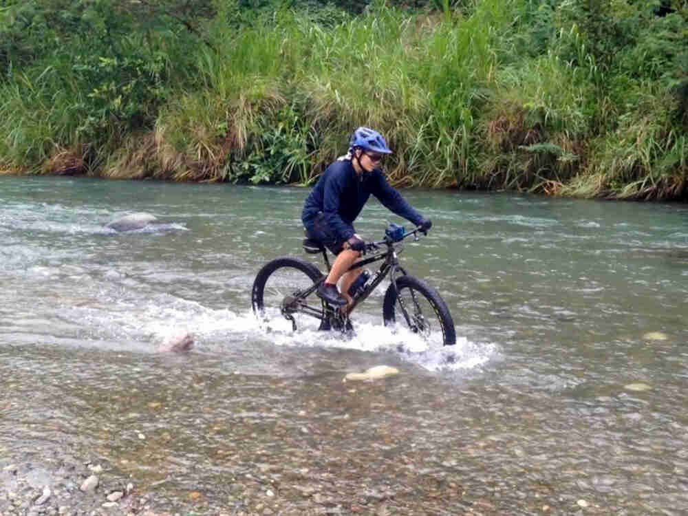 Right side view of a cyclist on a Surly Krampus bike, riding down a river, with a grassy bank in the background
