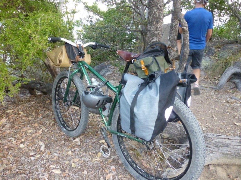 Rear, left side view of a green Surly Krampus bike with gear, parked on dirt, with a cyclist standing in the background