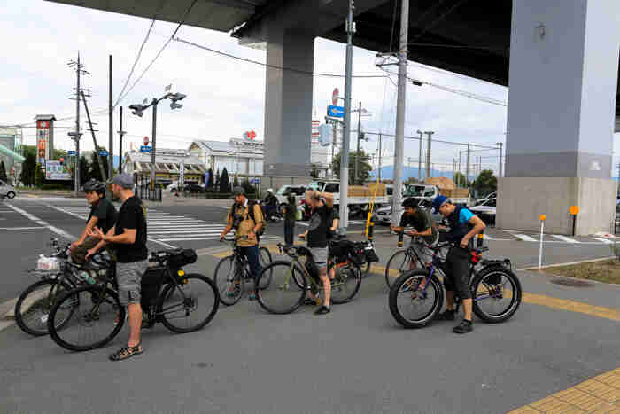 Left side view of a group of cyclists, standing over their bikes, parked underneath a cement, city bridge overpass