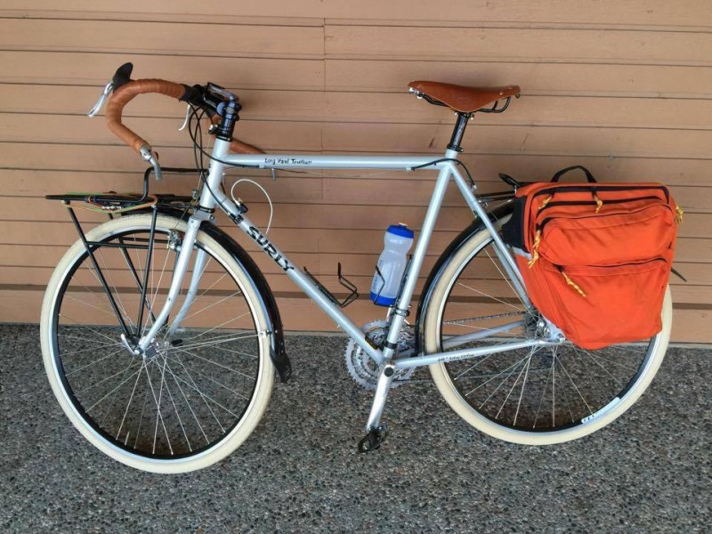 Left side view of a mint Surly Long Haul Trucker bike with white wall tires and rear saddlebags, parked against a wall