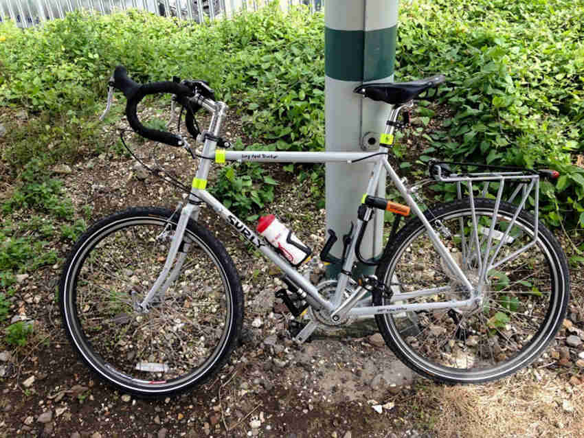 Left side view of a gray Surly Long Haul Trucker bike, parked on rocks in front of a steel post, with weeds behind