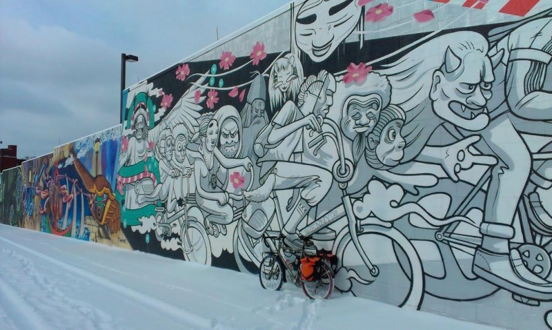Left side view of a Surly Long Haul Trucker bike with gear, parked against a long concrete wall with painted murals