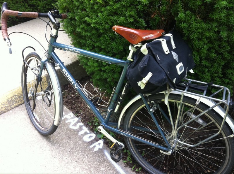 Downward, left side view of a bluish gray Surly Long Haul Trucker bike, parked on a sidewalk against a hedge