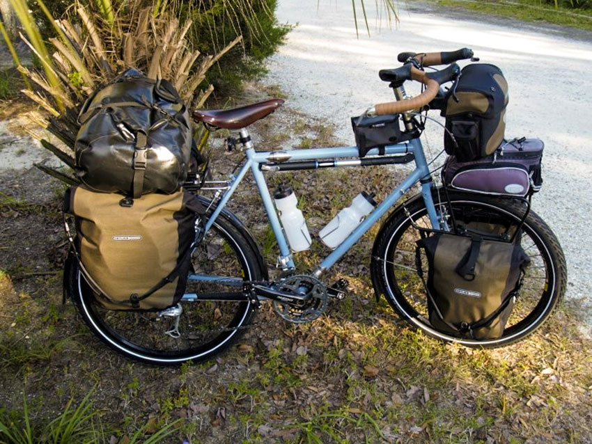 Right side view of a Surly Long Haul Trucker bike, blue, loaded with gear, parked in grass on the side of a gravel road