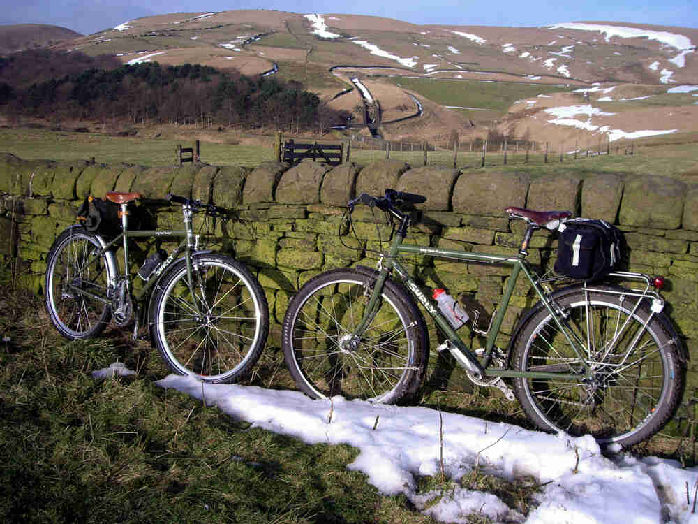 Side view of 2, olive green Surly Long Haul Trucker bikes, parked facing each other against a stone wall, with hills in the background