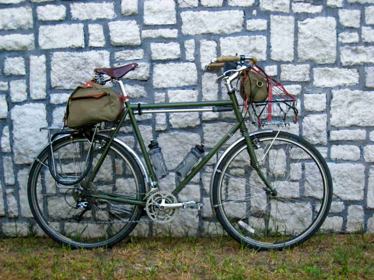Right side view of an olive green bike with front and rear rack packs, parked on grass and leaning against a stone wall