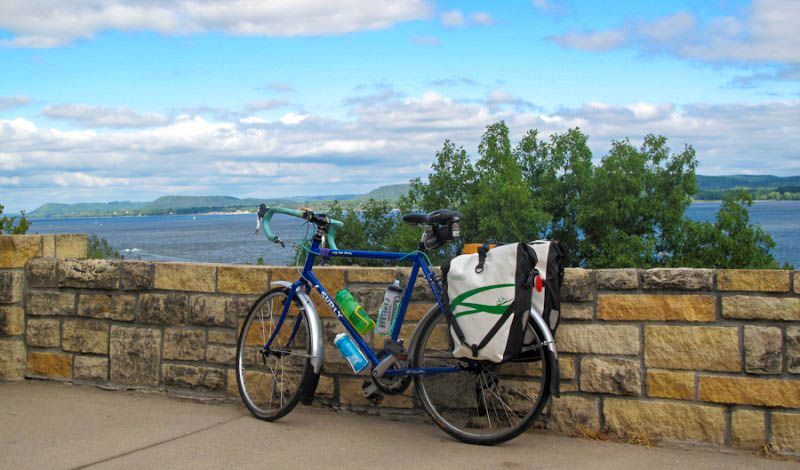 Left side view of a Surly Long Haul Trucker bike, parked on a sidewalk along a stone wall, with a lake in the background