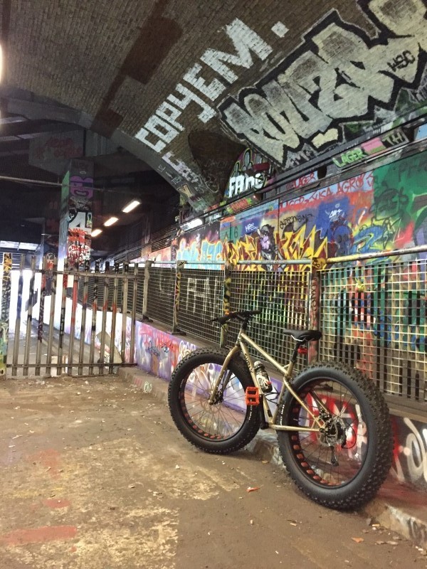 Left side view of a tan Surly Moonlander bike, parked against a railing, with a graffiti covered wall on the right