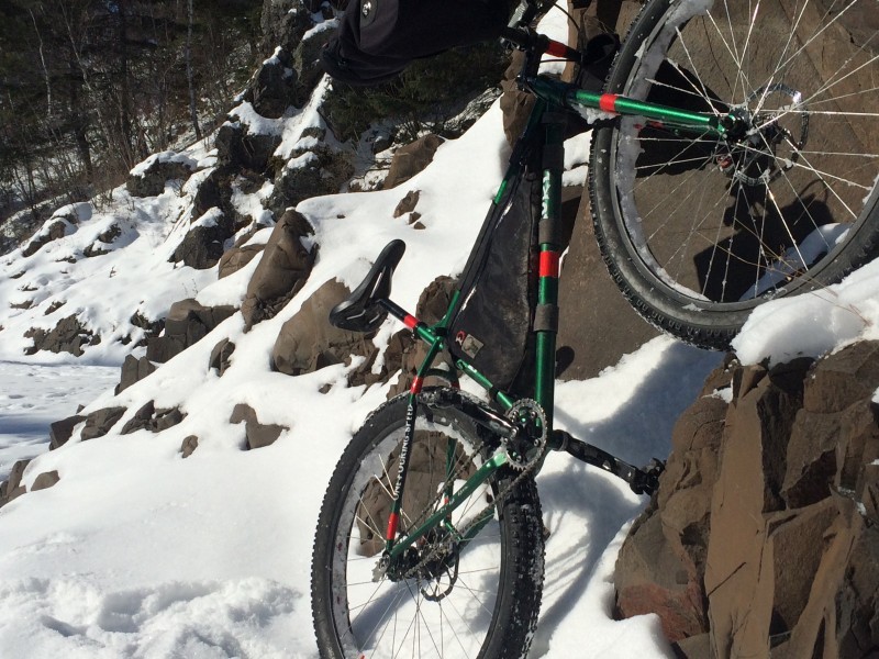 A Surly Krampus bike, green, right side view, facing upward on a large rock, at the base of a snowy cliff wall