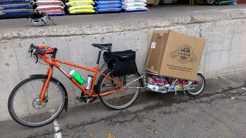 Left side view of an orange Surly Troll bike with rear saddlebags and loaded trailer, parked against a cement dock