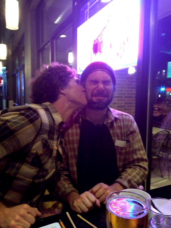 A person wearing a flannel shirt, kissing another person with a cringing face, on the check, while seated at a table