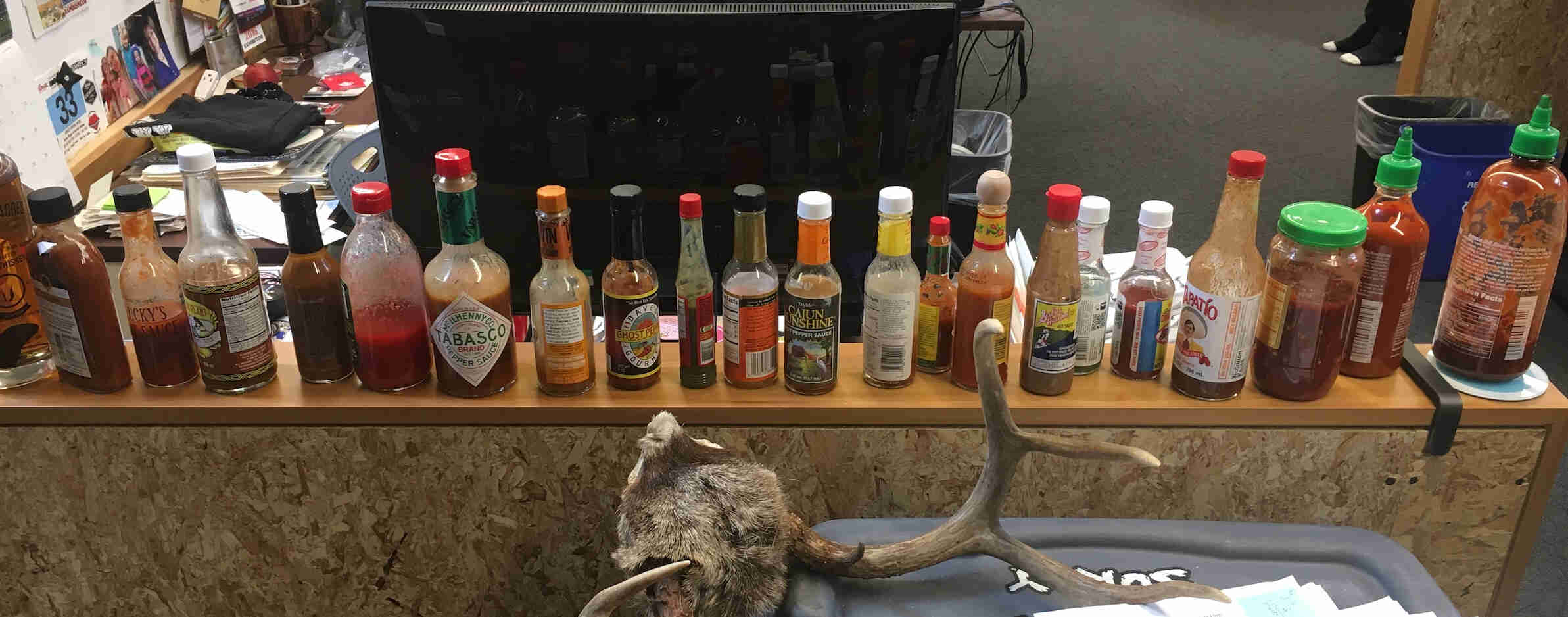 A row of hot sauce bottles lined up on a ledge, behind a tote with a deer antler on top