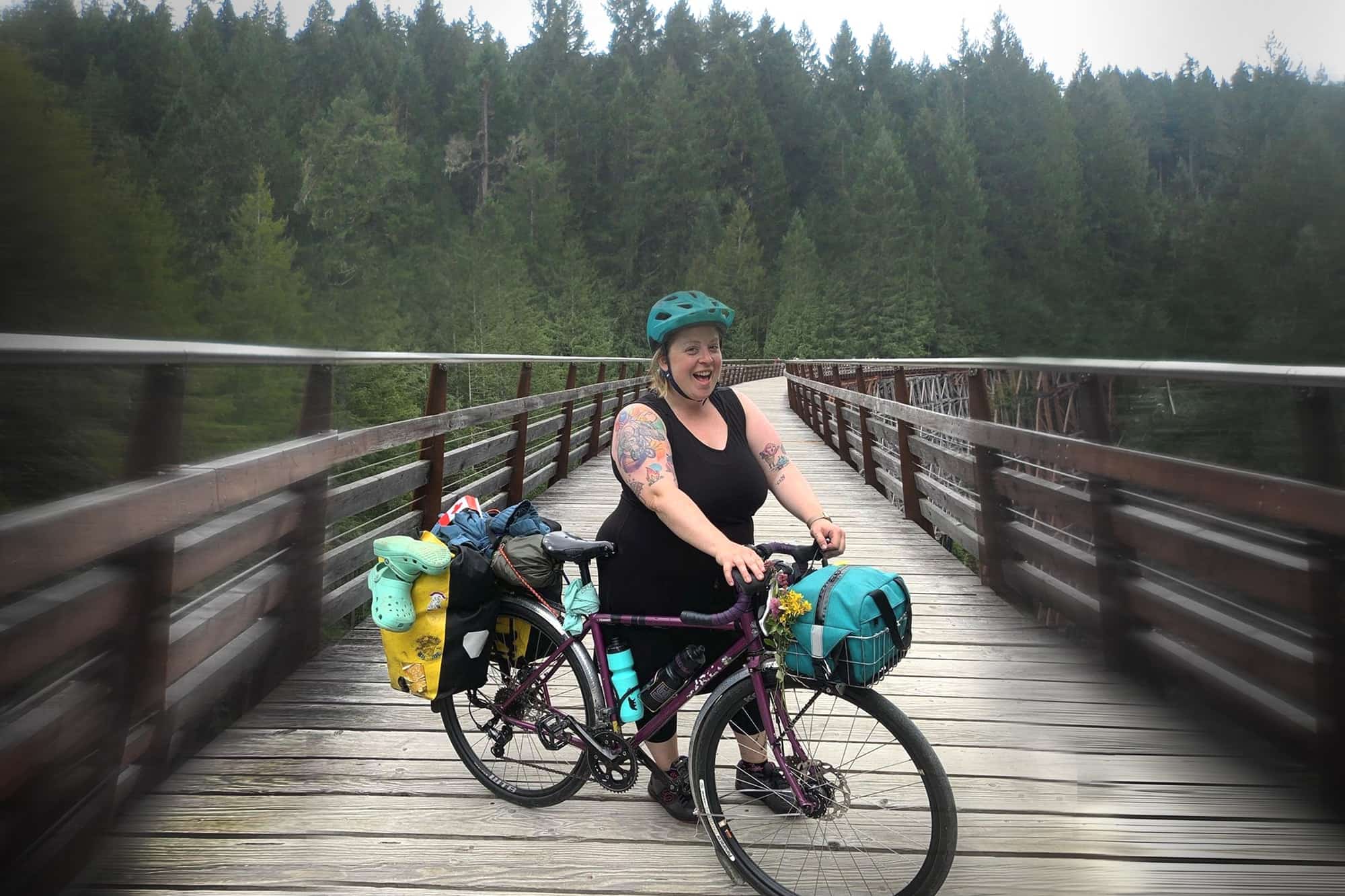 Cyclist stands on left side of fully geared Surly Straggler bike on wood bridge trail with pine trees in background