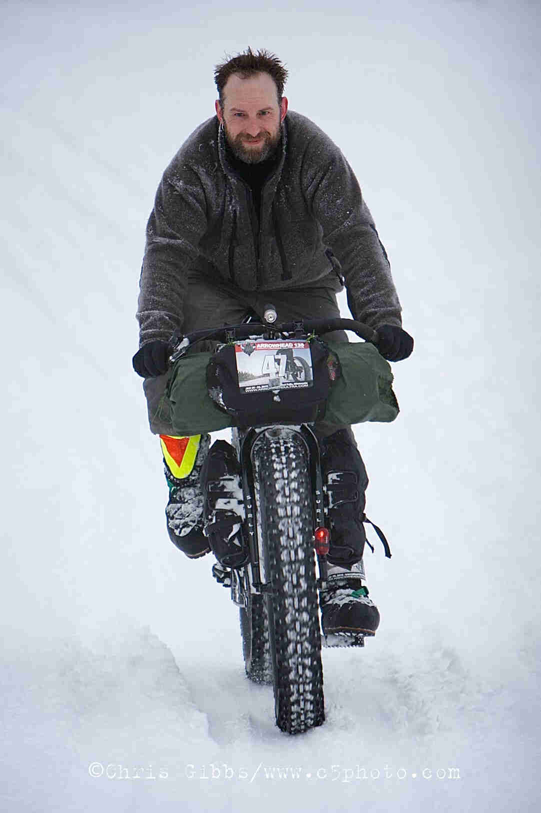Head on view of a cyclist, wear winter attire, riding a Surly fat bike loaded with gear, on a trail surrounded by snow