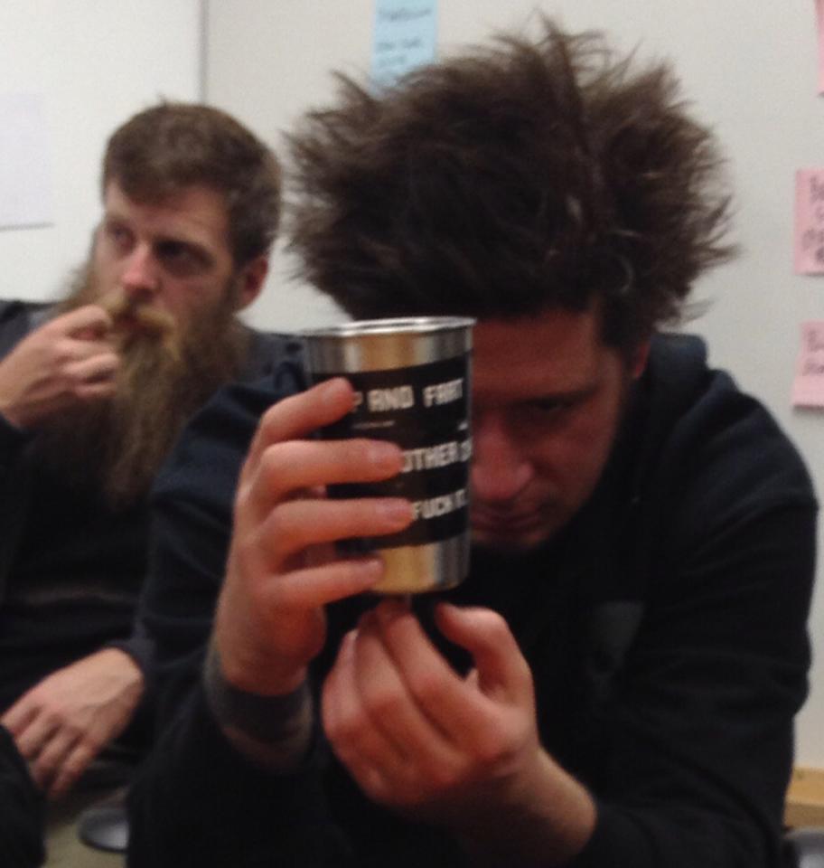 Front view of a person with bushy hair, holding a steel cup in front of their face, while sitting in a room