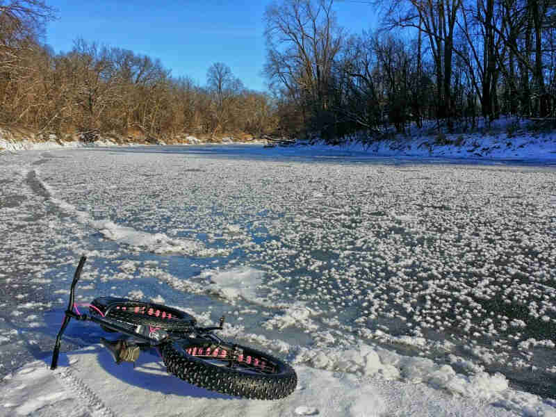 Rear view of a black Surly Moonlander fat bike, laying on it's left side, on a frozen river with bare trees on the sides
