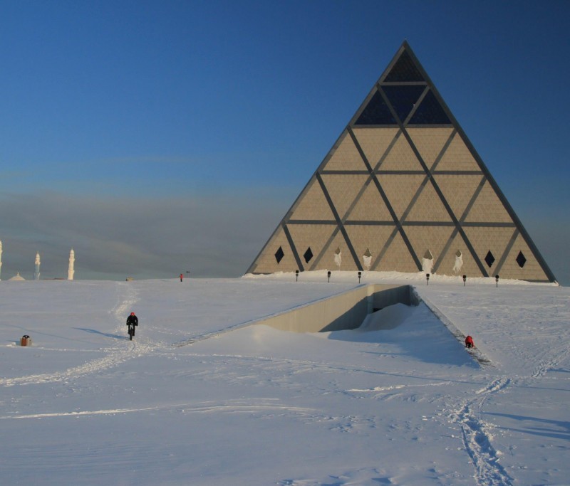 Front view of a cyclist, riding across a flat, snow covered area, with a pyramid building in the background